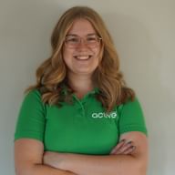 Leende blond kvinna med glasögon bär en grön Active pikétröja, står med korsade armar mot en vit vägg.