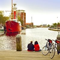 Två cyklister tar paus på träbrygga i Emdens hamn, sitter bredvid sina cyklar. Röda och vita fartyg förtöjda i bakgrunden.