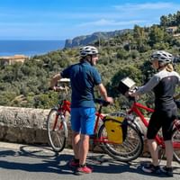 Två cyklister med hjälmar står bredvid röda turistcyklar med gula väskor på en kustväg på Mallorca med havsutsikt.