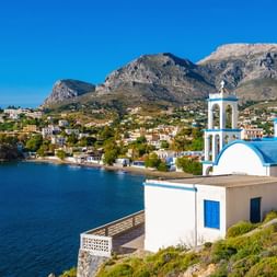 Vit ortodox kyrka med blå kupol och klocktorn på Kalymnos kulle med utsikt över azurblå vik med berg och vita byggnader.