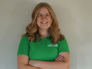 Leende blond kvinna med glasögon bär en grön Active pikétröja, står med korsade armar mot en vit vägg.