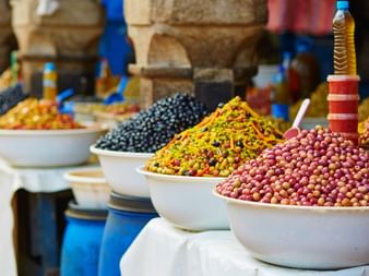 Vita keramikskålar fyllda med färgglada kryddor och oliver på en traditionell marockansk marknad i Essaouira, med stenbågar i bakgrunden.
