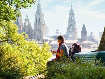 Kvinnlig pilgrim med ryggsäck sitter och läser på grön parkbänk, med utsikt över Santiago de Compostela katedralens torn.