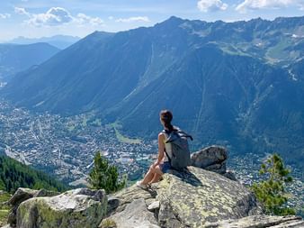 Vandrare med ryggsäck sitter på klippformation och blickar ut över fransk alpdal med stad nedanför, omgiven av dramatiska bergstoppar.