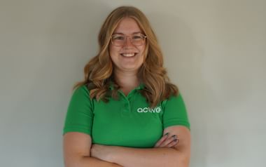 Mille från Active Scandinavia-teamet Leende blond kvinna med glasögon bär en grön Active pikétröja, står med korsade armar mot en vit vägg.