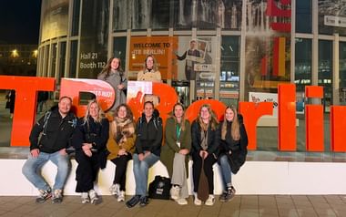 Grupp om tio personer poserar med stora orange ITB-bokstäver vid ingången till ITB Berlin. Skylt Hall 21 Monter 112 synlig i bakgrunden.