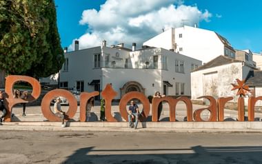 Stora orange metallbokstäver som stavar 'Sarramarin' med pilgrimer som sitter och vilar mot dem på ett stadstorg med vita byggnader.