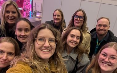 Gruppselfie med nio leende personer på resemässan ITB Berlin. Mässhall med vita väggar och rosa displayer i bakgrunden.