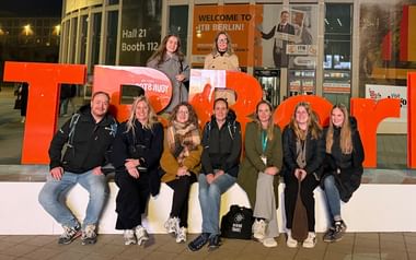 Grupp om tio personer poserar med stora orange ITB-bokstäver vid ingången till ITB Berlin. Skylt Hall 21 Monter 112 synlig i bakgrunden.