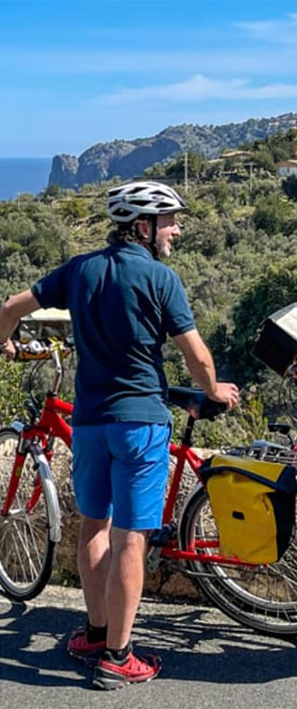 Två cyklister med hjälmar står bredvid röda turistcyklar med gula väskor på en kustväg på Mallorca med havsutsikt.