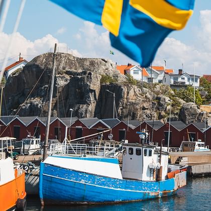 Blå och vit fiskebåt i Göteborgs skärgårdshamn med röda träbyggnader, klippor och svensk flagga i förgrunden.