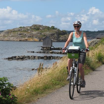 Female cyclist with helmet and green bike on coastal path along Kattegattleden. Rocky islands and small wooden pier visible in calm water.