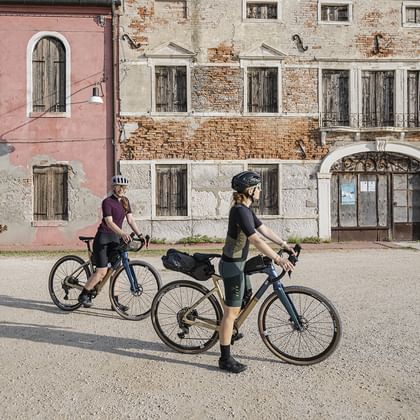 Två cyklister med turistcyklar står på ett soligt torg omgivet av historiska italienska byggnader med rosa och stenfasader.