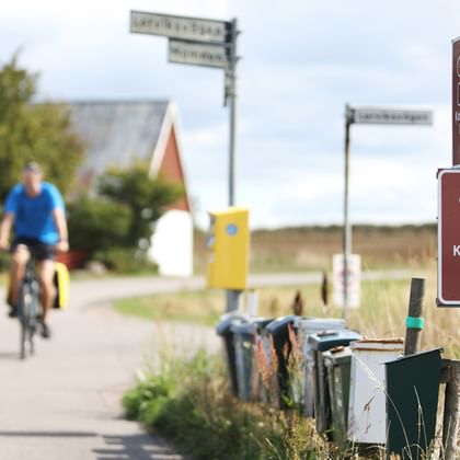 Brunt cykelrutt-skylt för Kattegattleden med cykelsymbol och pil. Två cyklister cyklar på vägen i bakgrunden förbi hus med röda tak.
