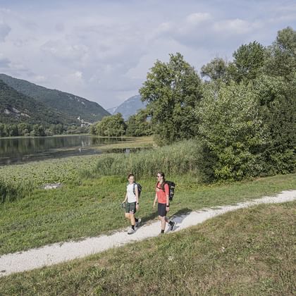 Två vandrare går på en grusväg bredvid en lugn sjö omgiven av gröna kullar och träd i den italienska Prosecco-regionen.