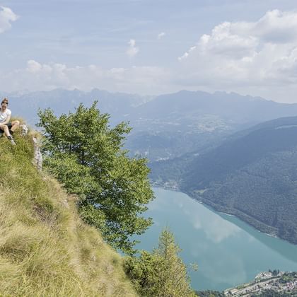Två vandrare sitter på en gräsbevuxen bergssluttning med utsikt över en turkos sjö omgiven av skogklädda berg.