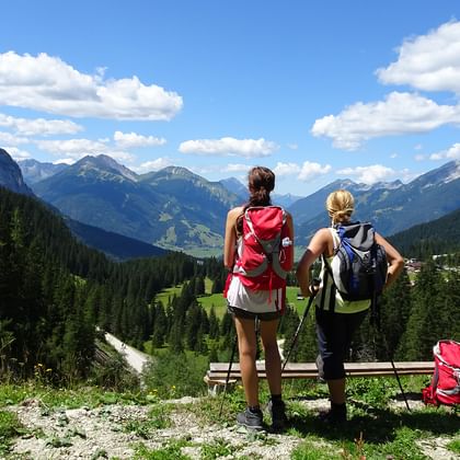 Två vandrare med ryggsäckar står vid träräcke och blickar ut över tysk alpdal med skogklädda berg och blå himmel med vita moln.