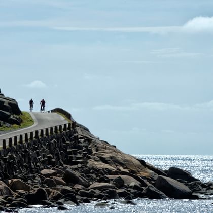 Two cyclists on curved coastal road with stone barriers along rocky shoreline. Sparkling sea and cloudy sky visible in background.