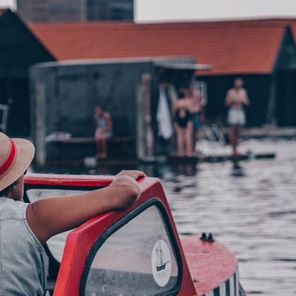 Man in straw hat with red band steering a red boat in Copenhagen harbor, with historic waterfront buildings and people visible in background.