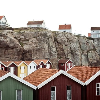 Bunte schwedische Sommerhäuser mit roten Ziegeldächern am Fuß dramatischer Felsklippen. Moderne weiße Häuser sind oben auf den Klippen sichtbar.