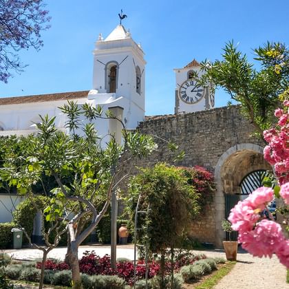 Vit Igreja do Castelo kyrka i Tavira med klocktorn och urtorn, omgiven av rosa blommande träd och trädgårdsvägar under blå himmel.