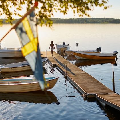 Wooden dock with moored boats on calm lake water during golden hour. Person swimming near the pier with Swedish flag visible on boat.